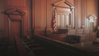 An empty court room with wood-paneled walls, with a judge's bench and witness stand in view.