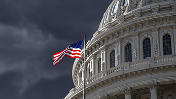 An American flag waves in front of the Capitol with a stormy sky