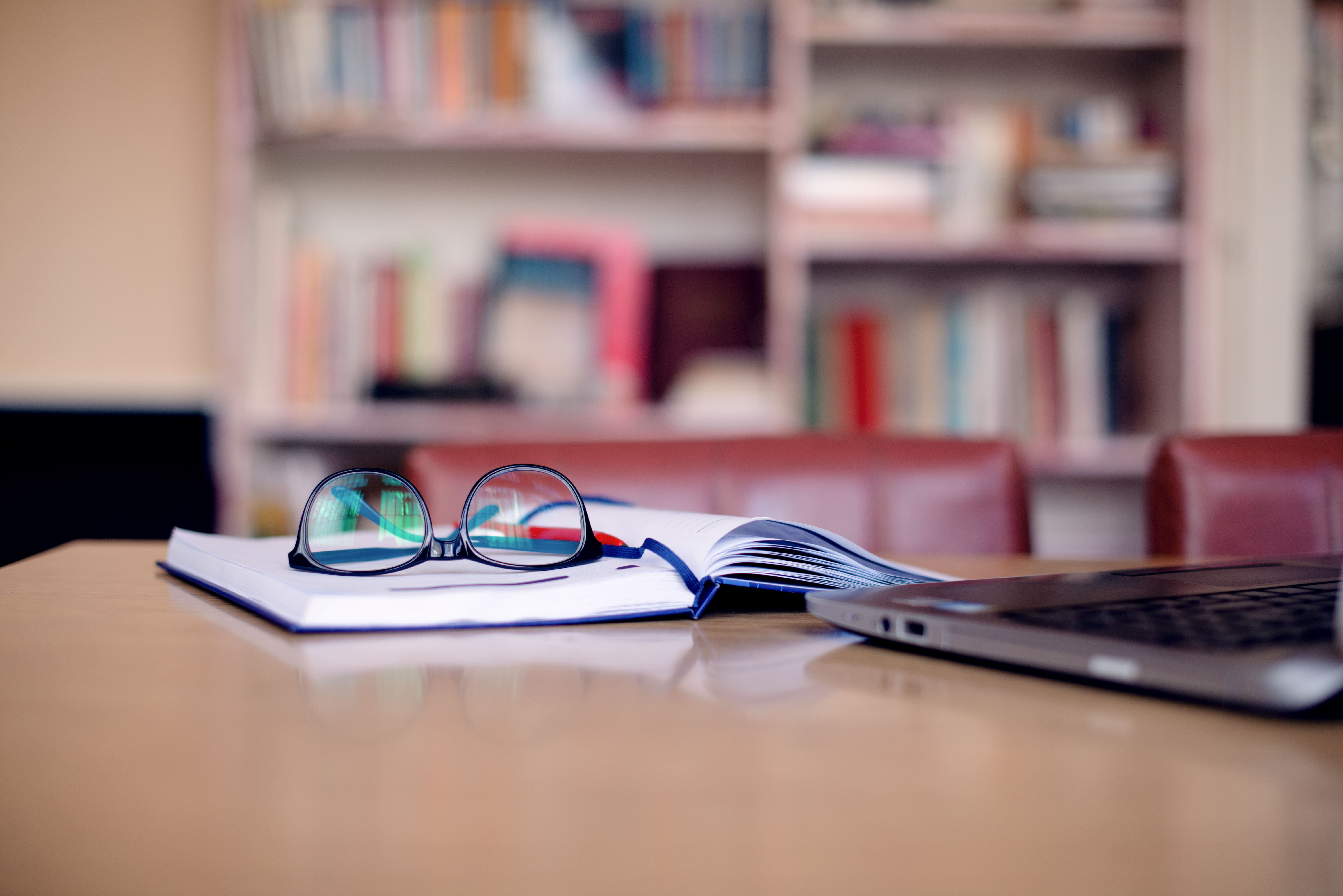 Reading glasses on top of an open book with a laptop on top of a desk
